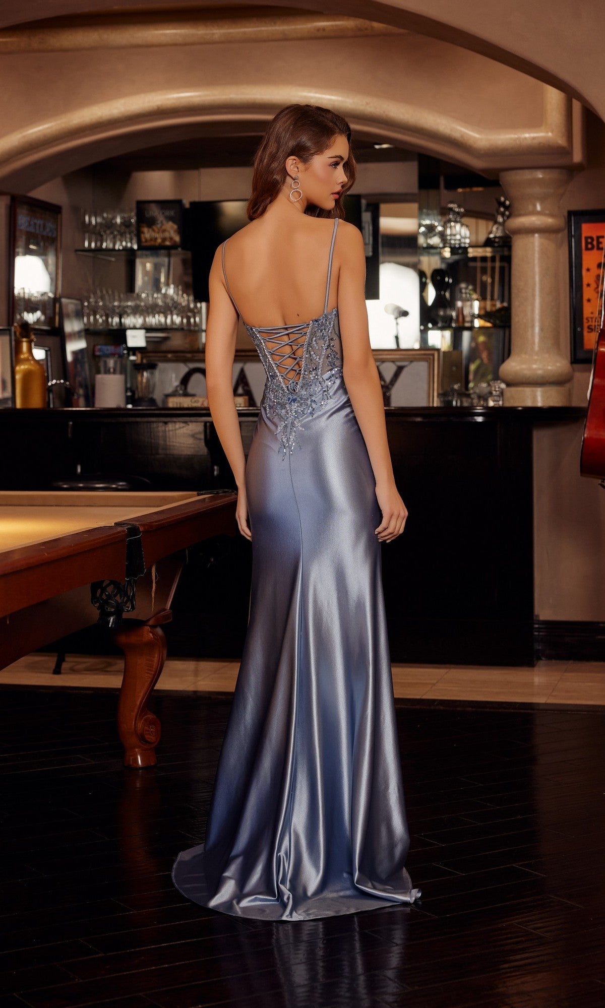 A woman stands near a billiards table in a bar or lounge, facing away from the camera, wearing the Nox Anabel D1766 long prom dress with an embellished corset bodice, sleeveless design, and lace-up back.
